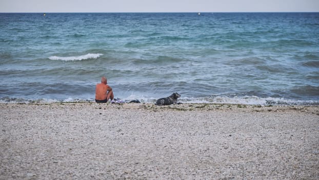 Elderly man sitting on pebble beach with his dog, enjoying the view of the Black Sea in Crimea