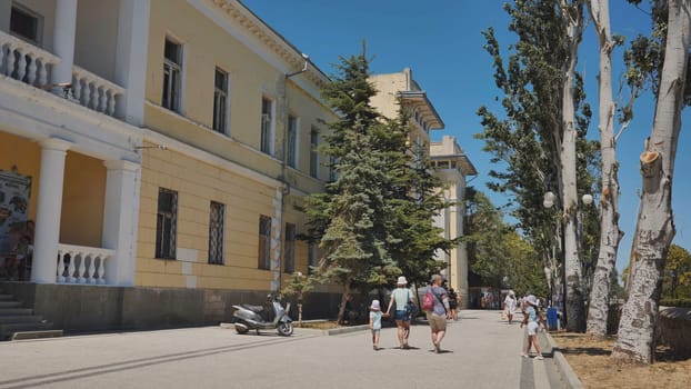 Tourists walking sunny promenade, lined with trees, historic architecture framing scenic waterfront landscape in crimean coastal setting