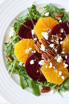 A vibrant salad features fresh arugula topped with orange slices, roasted beets, crumbled cheese, and toasted pecans. A drizzle of dressing completes the dish presented on a white plate.
