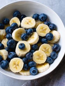 A bowl filled with sliced bananas and fresh blueberries sits on a gray surface. This nutritious combination offers a vibrant look and a delicious flavor, perfect for a healthy snack.