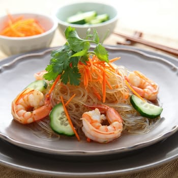 A vibrant dish featuring translucent noodles topped with shrimp, fresh cucumber slices, and colorful carrot shreds. Two small bowls of dipping sauce accompany the plate, ideal for a light meal.