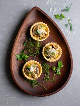 Three savory tarts filled with mushrooms, herbs, and cheese are elegantly arranged on a wooden platter. Fresh herbs are scattered around, enhancing the presentation.