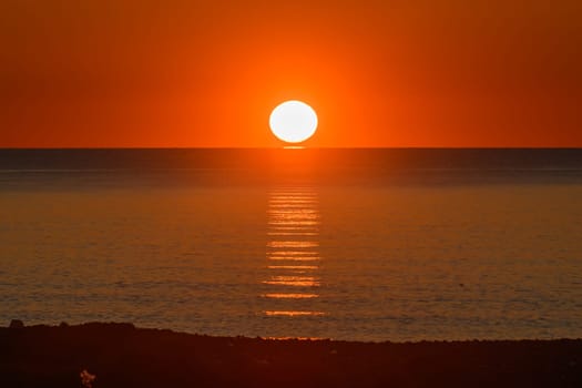 Vibrant hues of orange and gold paint the sky as the sun dips below the horizon, casting shimmering reflections on the calm ocean surface, creating a tranquil and magical atmosphere.