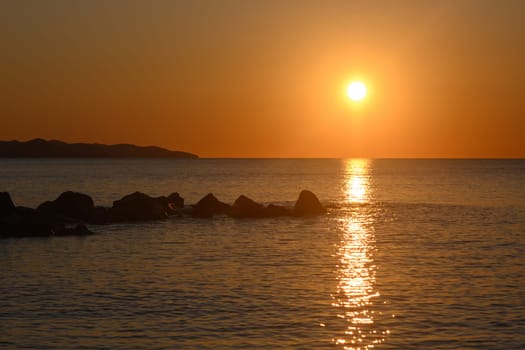 The sun slowly descends in a fiery display, casting warm orange and yellow hues over the serene ocean. Waves gently lap against dark rocks, creating a peaceful ambiance as twilight approaches.