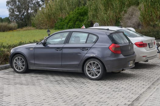 Gaziveren Cyprus -10.04.2025 In a peaceful parking area, a dark gray car and a light-colored vehicle sit side by side. The backdrop features gentle greenery under an overcast sky, offering a serene atmosphere.
