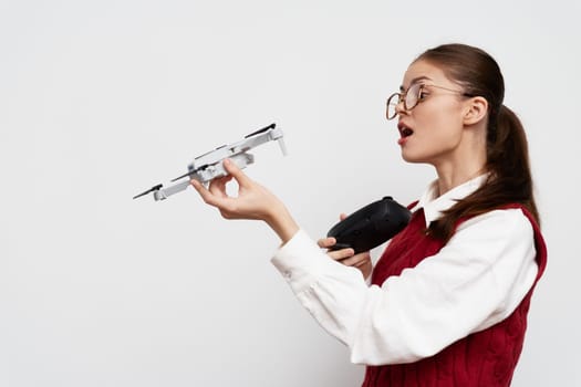 A young woman with glasses is holding a small drone in one hand and a remote control in the other, showcasing a modern indoor setting with a white background, expressing focus and curiosity about tec