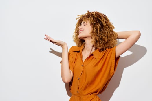 A cheerful woman with voluminous curly hair is showcasing a playful pose against a clean, light colored background Dressed in an orange outfit, she radiates warmth and positivity