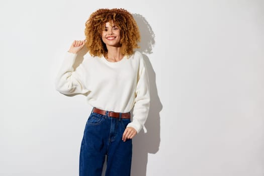 cheerful woman with voluminous curly hair dressed in a white sweater and blue denim jeans, standing against a plain white backdrop, exuding confidence and friendliness