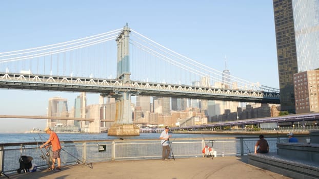 New York City, United States - 7 Sept 2023: Manhattan Bridge. Fisherman near Pier 35 swings, waterfront downtown skyline cityscape. World Trade Center skyscraper, people fishing and morning sunset.