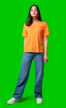 A model stands upright wearing a bright orange shirt and blue jeans, showcasing a confident expression. The vivid green backdrop highlights the subject during a studio session.