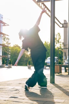 A dancer engages in expressive movement in an urban area as the sun sets, casting a beautiful glow and creating a striking silhouette against the backdrop of trees and buildings.