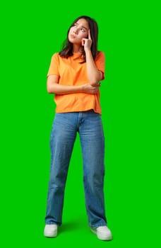 A young woman stands with her hand on her chin in a thoughtful pose. She wears an orange shirt and blue jeans, contemplating ideas in a vibrant setting.