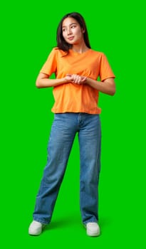 A young woman stands with her hands clasped, wearing an orange shirt and blue jeans. The vibrant green background highlights her relaxed yet confident pose.