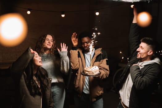 Friends are gathered in a warm environment, joyfully celebrating an occasion. They are raising their hands in excitement while a cake with sparklers is presented, creating a festive atmosphere.