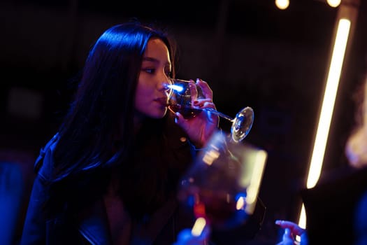 A woman with long dark hair sips from a glass in an atmospheric setting. The venue features vibrant lighting, creating a lively backdrop for her evening out with friends or acquaintances.