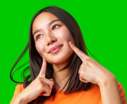 A young woman smiles while pointing at her cheeks, showcasing a playful demeanor. She stands in front of a bright green background, wearing an orange shirt, capturing a joyful moment.