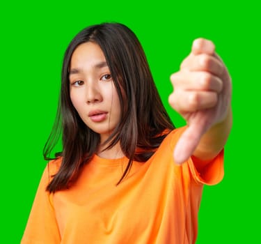 A young woman wearing an orange shirt displays a thumbs down gesture. Her expression conveys disapproval or disappointment, emphasizing her message clearly against a vivid green backdrop.