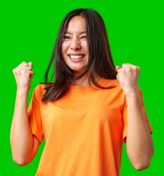 A young woman wearing a vibrant orange shirt is joyfully expressing her excitement, with her fists raised in celebration. The bright green background enhances the lively atmosphere.