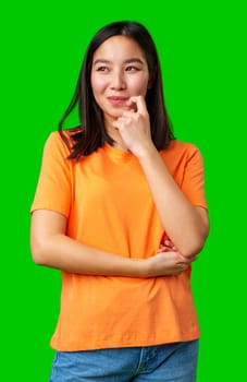 A young woman stands with a contemplative expression, gently biting her finger. She wears a vibrant orange shirt and has long, dark hair, exuding a casual, relaxed vibe.