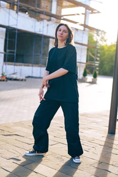 A young woman wearing a loose black shirt and wide-legged pants strikes a pose in front of a construction site, basking in the warm light of the setting sun.