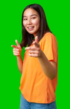 A young woman smiles confidently while playfully posing with finger guns. She wears a bright orange shirt, adding a lively contrast to the vibrant green background.