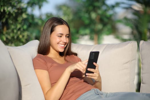 Happy woman using mobile phone sitting in sofa in a terrace at home a sunny day