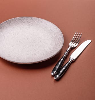 Empty ceramic plate on a monochrome brown background. Empty plate with cutlery on a dining table