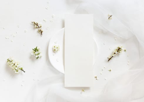 Blank menu card on plate near tulle and wild plums white flowers top view, mockup. Pastel vertical card for spring party design, mothers day or romantic wedding stationery