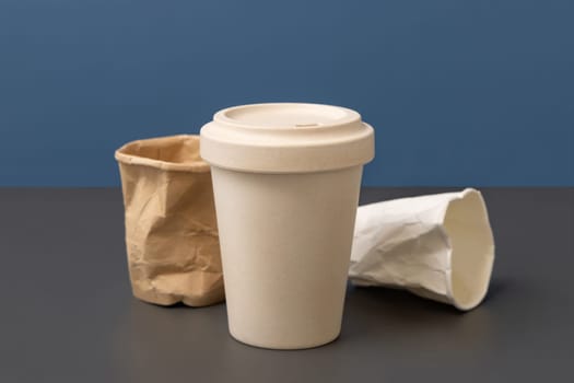Used and crumpled disposable paper cups and reusable bamboo coffee cup on gray against blue closeup, copy space , mockup. Consumerism concept. No plastic and sustainable lifestyle concept