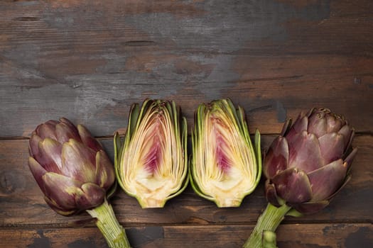 Half and whole purple raw roman artichokes on board on dark weathered wooden table top view, copy space. Fresh vegetables bought on farmers market ready to cook. Italian cousine ingredient
