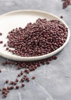 Ceramic plate full of dried red azuki beans on gray table closeup, copy space. Healthy vegetarian food, legumes protein source. Traditional East Asiancousin ingredient