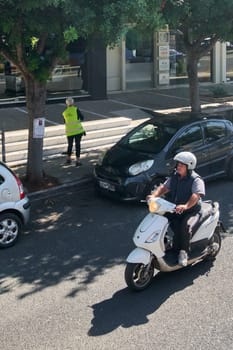 Crete.Greece - april 14, 2025: A motorcyclist wearing a helmet rides past a municipal worker in a high-visibility vest sweeping the sidewalk beside parked cars and a tree.