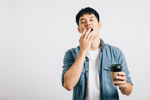 A young man, overwhelmed by fatigue, yawns with his eyes closed, coffee cup in hand. Studio shot isolated on white, his tired expression tells a relatable story. Copyspace included.