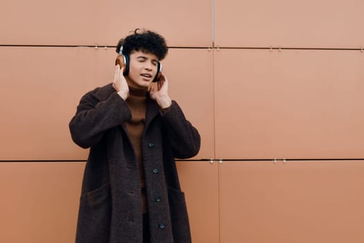 Fashionable asian man listening to music with headphones, standing in front of an orange wall. trendy stock photo of a young man enjoying music.