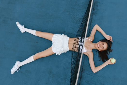 Joyful young woman in white athletic attire, relaxing on a blue tennis court, holding a tennis ball, exuding confidence and positivity in a vibrant sports setting