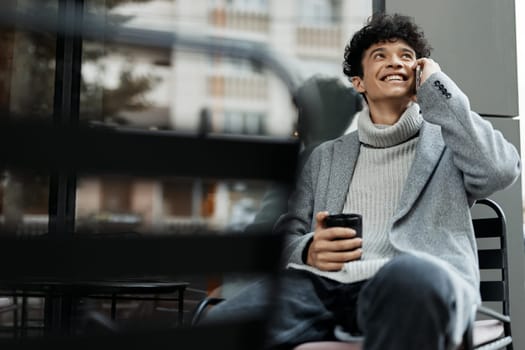 Fashionable man sitting on bench, talking on phone and holding coffee cup in front of face while looking at camera. trendy lifestyle concept.