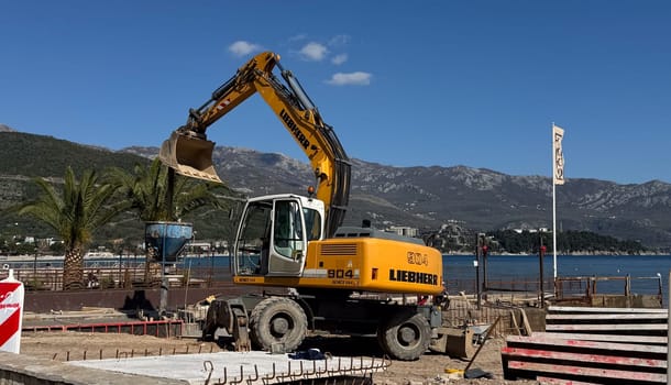 Budva,Montenegro-April 11,2025:A yellow Liebherr excavator working near the coast filling a concrete crater at a coastal construction site with palm trees and a COCO cafe flag. High quality photo