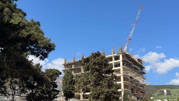High rise concrete structure under construction with scaffolding and tall red and white crane against clear blue sky.
