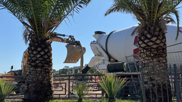 Budva,Montenegro-April 11,2025:Construction equipment including excavator bucket and concrete mixer on the shore next to a cafe surrounded by palm trees and metal fence. High quality photo