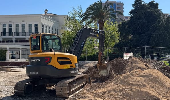 Budva,Montenegro-April 11,2025: Yellow and black Volvo excavator digging soil at a city construction site surrounded by buildings and palm trees.