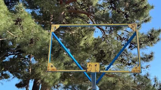 A rusty yellow and blue metal basketball hoop stands against a background of pine trees, without a net or backboard, evoking a sense of antiquity. ... High quality photos