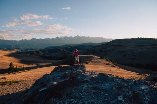 A woman in casual clothing taking photo on her smartphone on a rocky peak, gazing at scenic mountains and a vast valley under golden sunlight.
