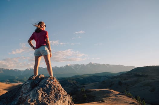 A woman in casual clothing stands on a rocky peak, gazing at scenic mountains and a vast valley under golden sunlight.