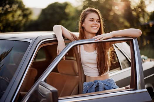 A cheerful young woman in a white tank top and denim shorts leans against her car, exuding joy and relaxation on a sunny day, symbolizing freedom and outdoor adventures.