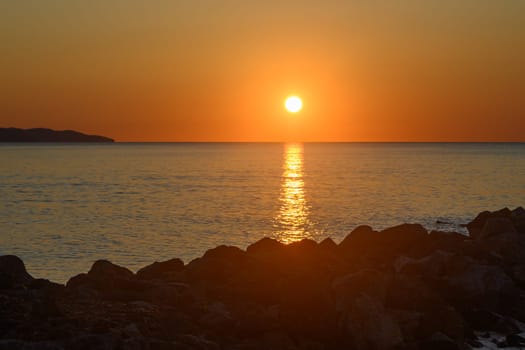 As the sun sets on the horizon, hues of gold and orange illuminate the calm sea, creating a peaceful ambiance. Silhouetted rocks frame this picturesque moment, inviting tranquility.