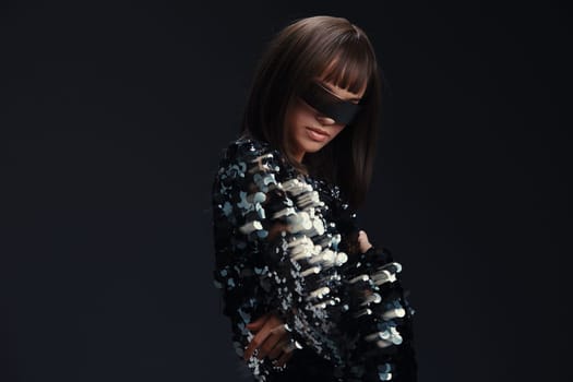 Woman in black and white dress, blindfolded, wearing necklace and head scarf, posing against neutral background, evoking themes of mystery, fashion, and intrigue