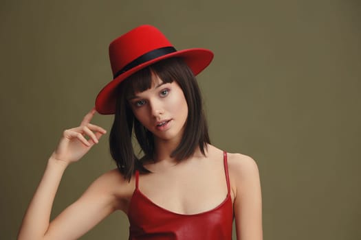 Woman, red hat, leather dress, green background, hand on head, finger on lips a stylish woman in a red hat and leather dress poses against a green backdrop, with one hand on her head and a finger on