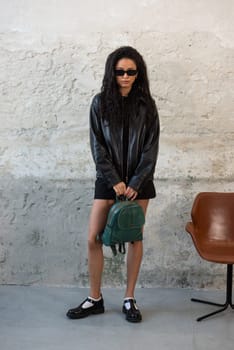 Curly girl wearing black leather jacket, sunglasses, shoes and short skirt posing with a green backpack.