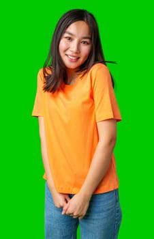 A young woman with long hair smiles and poses playfully while wearing an orange shirt and jeans. The vibrant green background adds energy to her joyful expression.