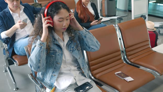 Travel vacation. Happy Asian young woman curly hair listening music with headphone and mobile phone watching something on social media at airport terminal hall while waiting for her flight